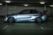 Side view of a BMW M2 sports car with racing stripes, parked in a dimly lit garage.